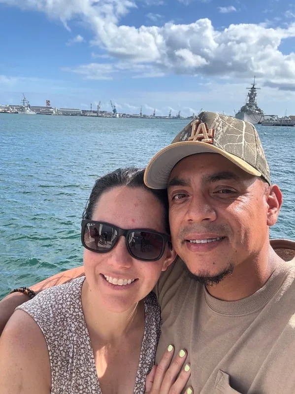 Smiling couple posing for a selfie by the water with ships and a blue sky with clouds in the background.