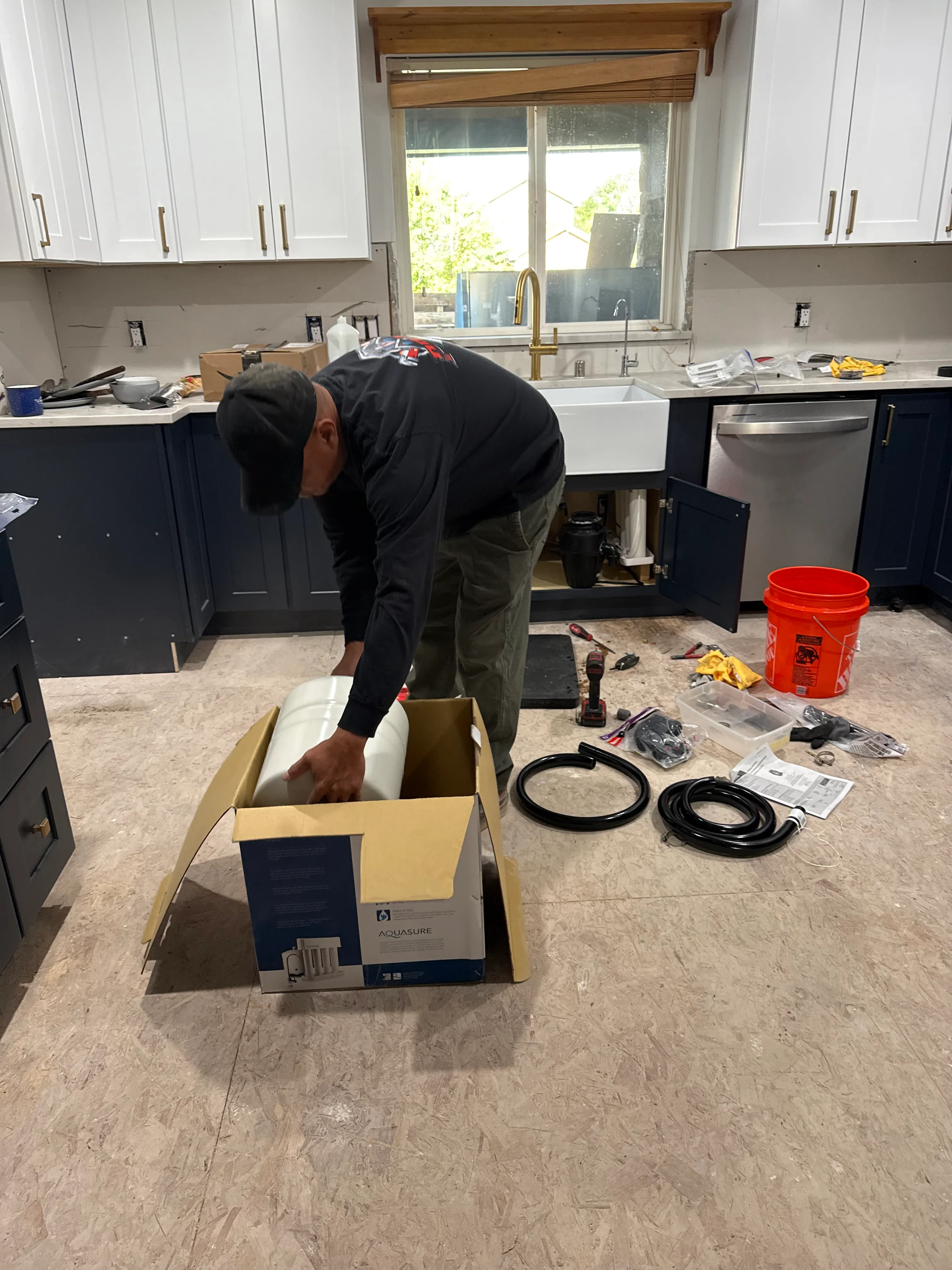 Person installing plumbing equipment in a kitchen under a window with white and navy cabinets and tools scattered on the floor.