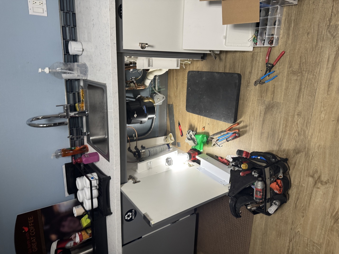 Open kitchen cabinet below a sink with plumbing exposed, surrounded by various plumber tools on a wooden floor.