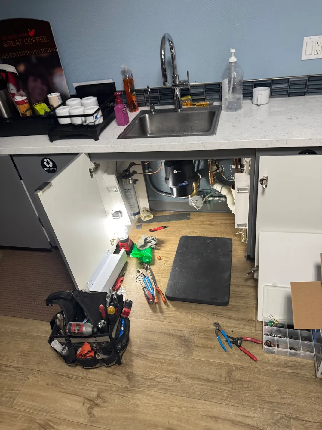 Open kitchen cabinet below a sink with plumbing exposed, surrounded by various plumber tools on a wooden floor.