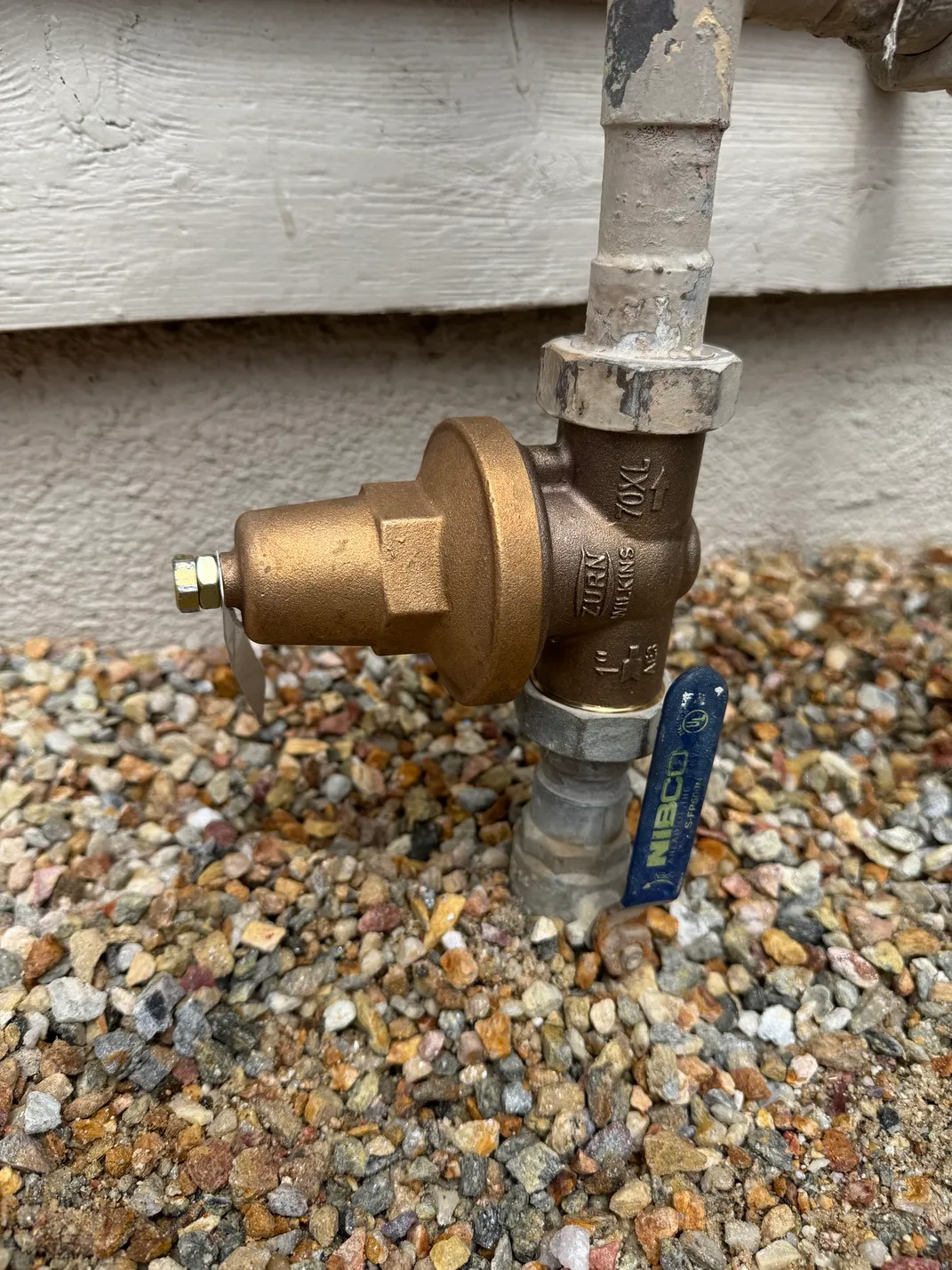 Close-up of a bronze water valve connected to a pipe, mounted near a gravel ground and a white wooden wall.