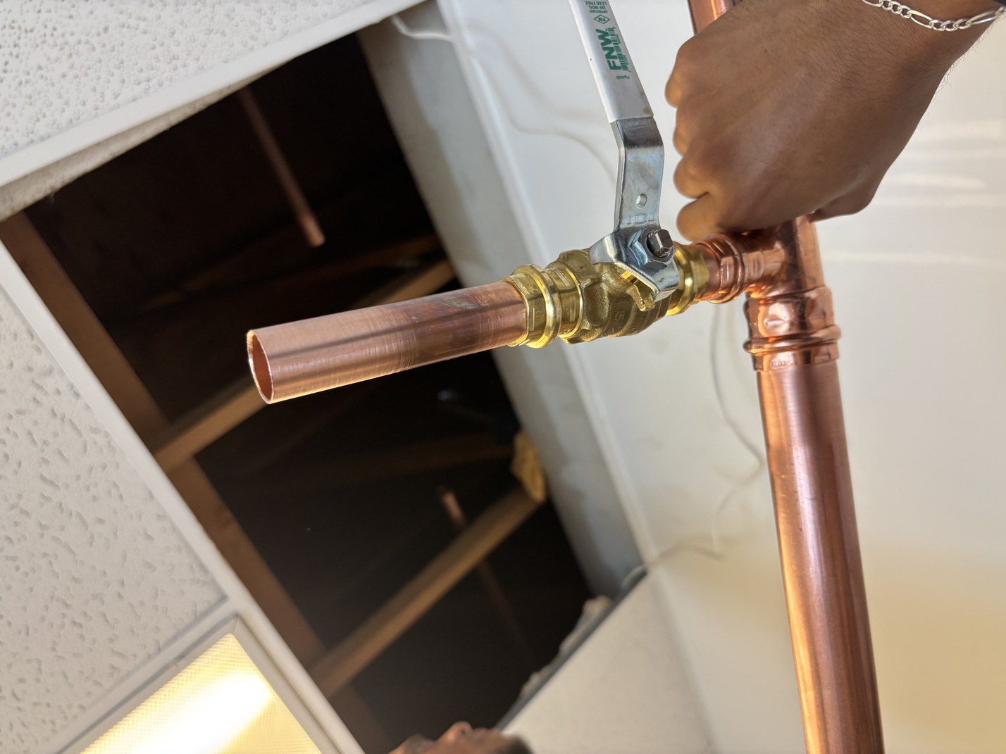 Hand holding a copper pipe with a brass ball valve in a ceiling panel opening.