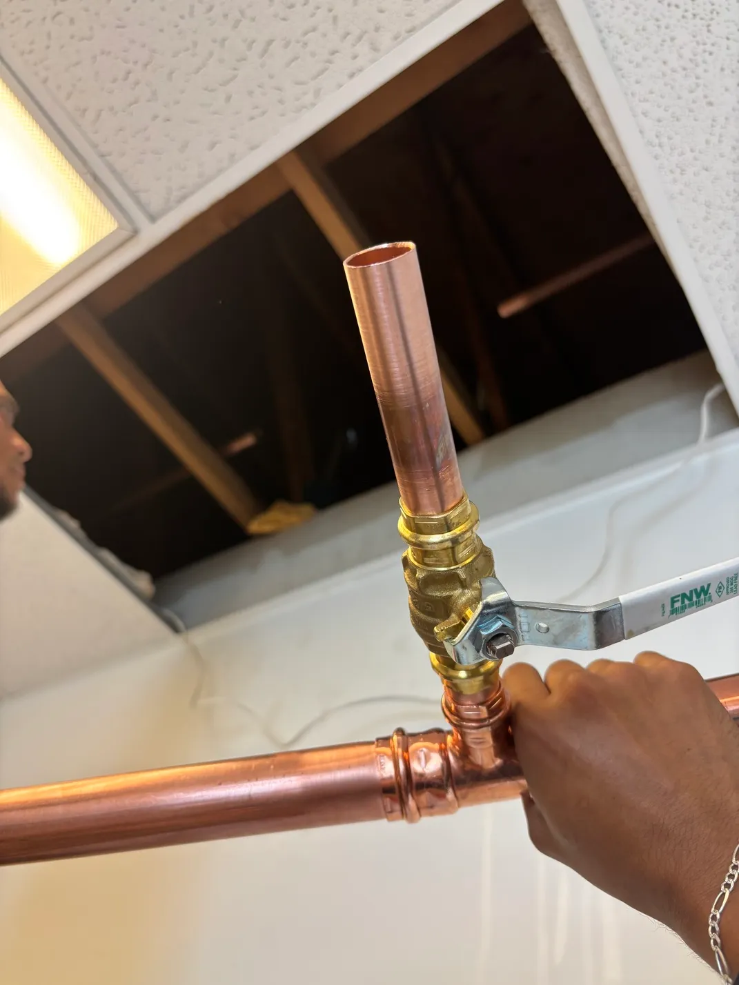 Hand holding a copper pipe with a brass ball valve in a ceiling panel opening.