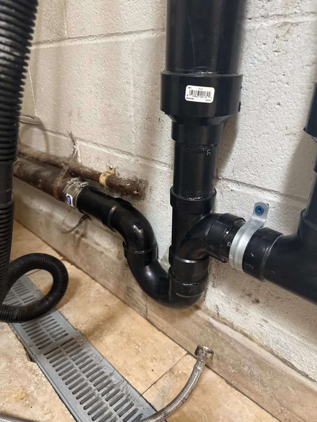 Close-up of black PVC drain pipes connected with a coupling, mounted on a cinder block wall above a floor drain.