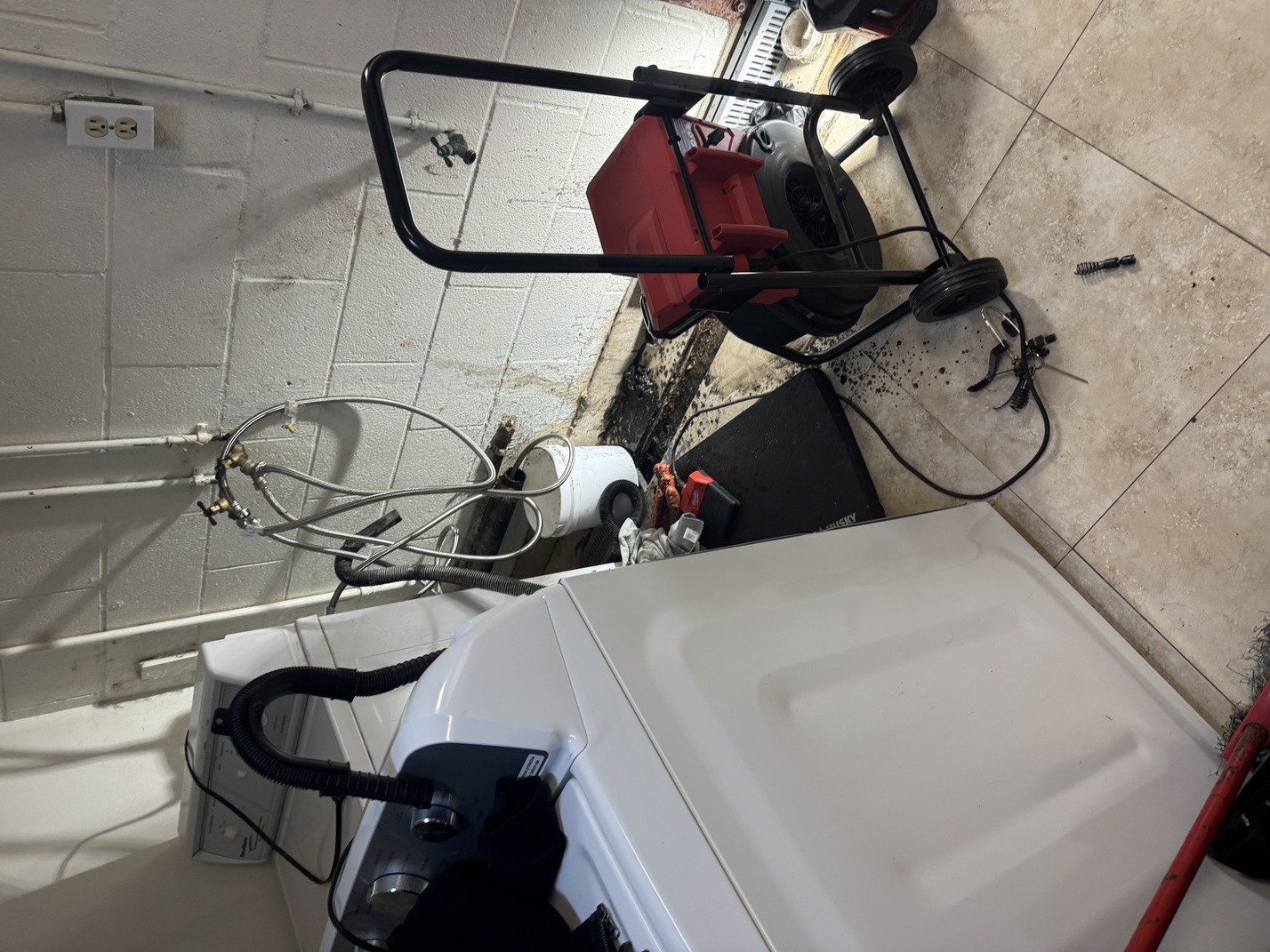 Laundry room corner with a washing machine, wall plumbing hookups, a bucket, and a drain cleaning machine on wheels with attached cables and tools on tiled floor.
