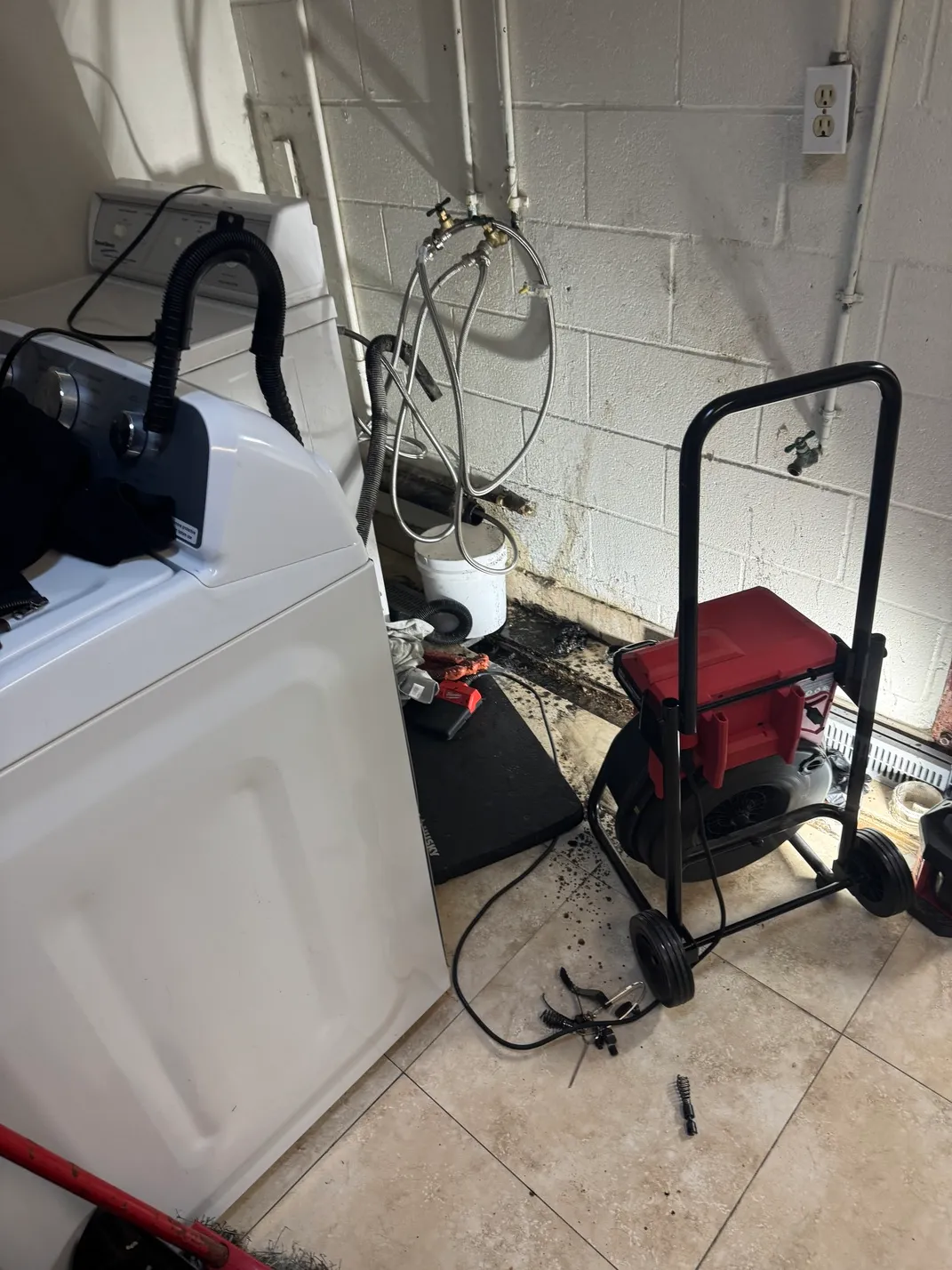 Laundry room with a white washing machine, cleaning equipment including a black and red drain cleaning machine on wheels, various hoses, and tools scattered on the tiled floor near a stained wall.