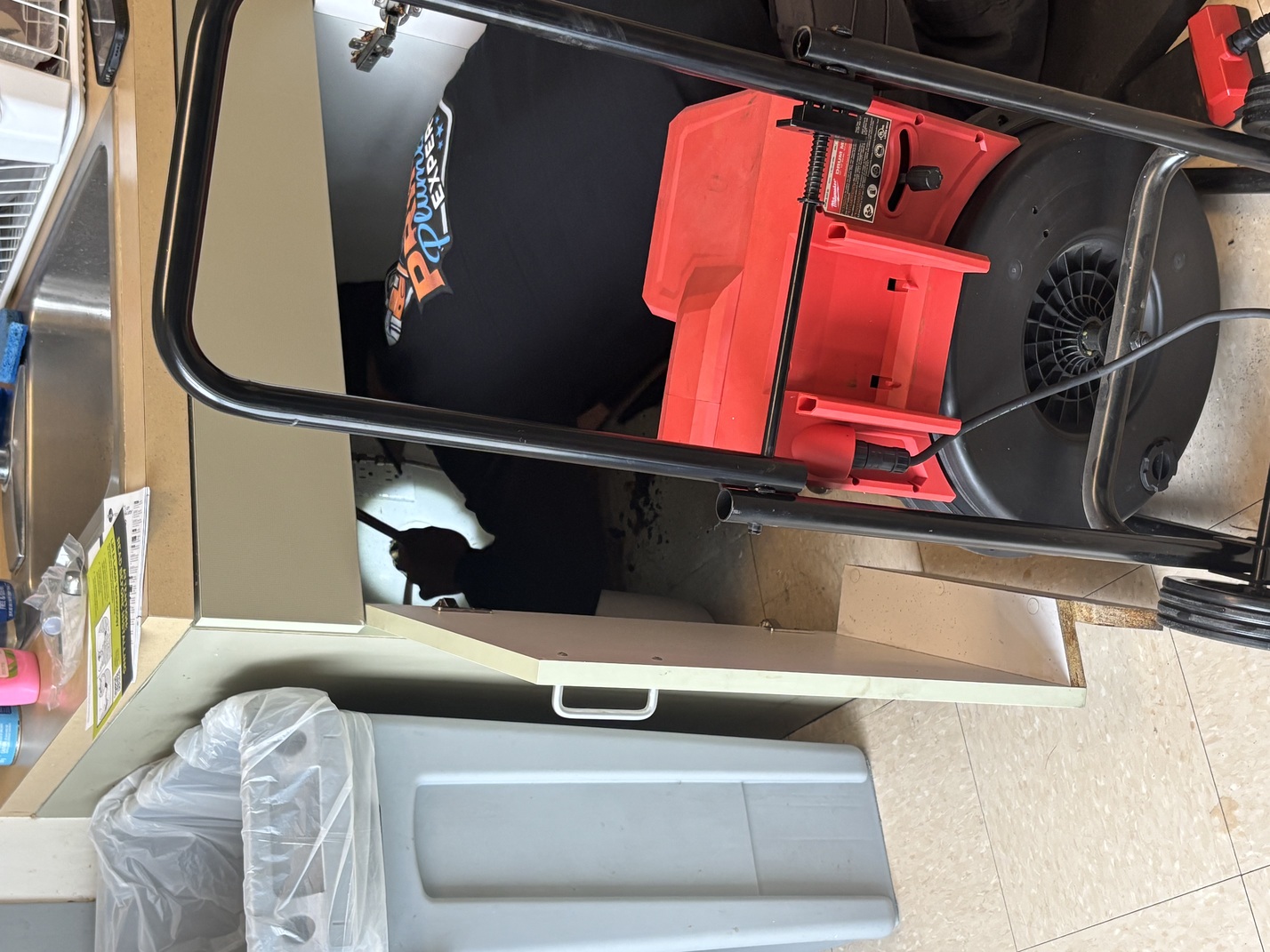 Plumber working under a kitchen sink with tools and a red drain cleaning machine in front.