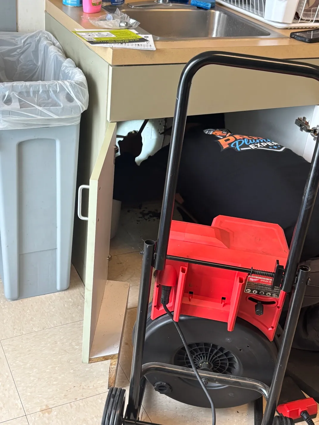 Plumber working under a kitchen sink with tools and a red drain cleaning machine in front.