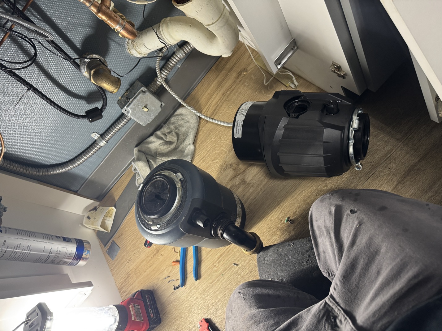 Under-sink workspace showing a removed garbage disposal, tools, pipes, and a worker's knee.