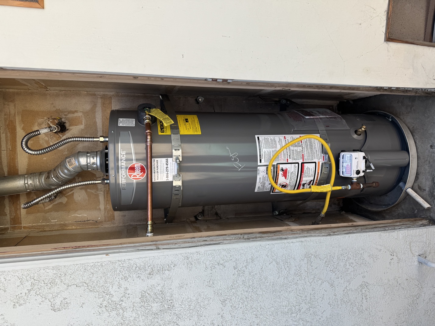 Gray Rheem Performance water heater installed in a small outdoor alcove with pipes and yellow gas line attached.