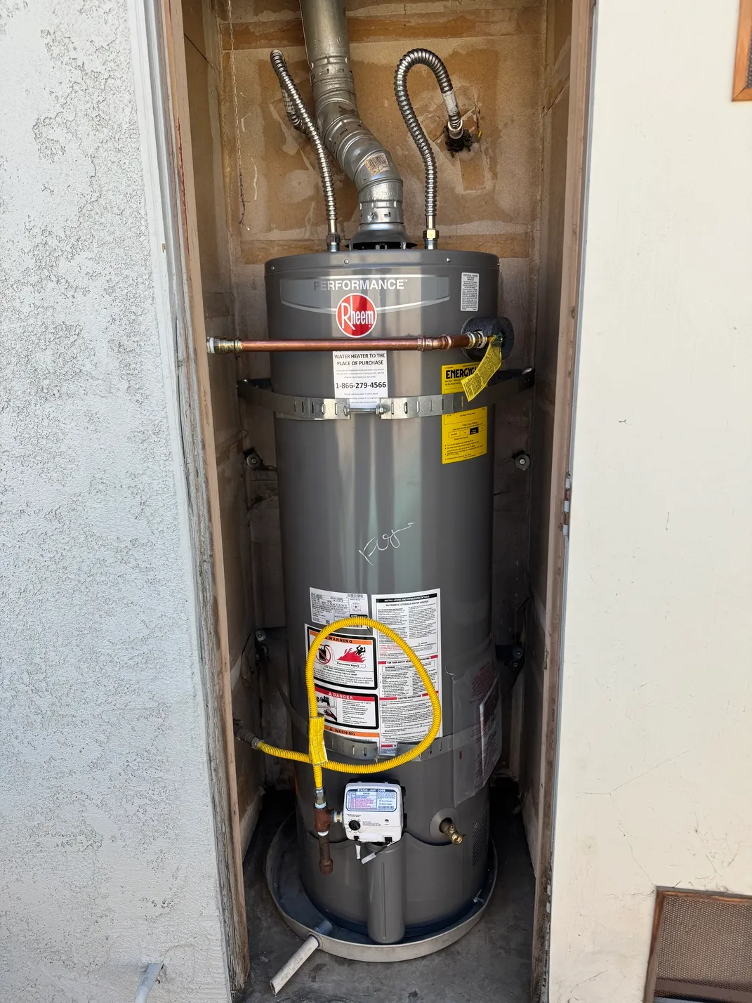 Gray Rheem Performance water heater installed in a small outdoor alcove with pipes and yellow gas line attached.