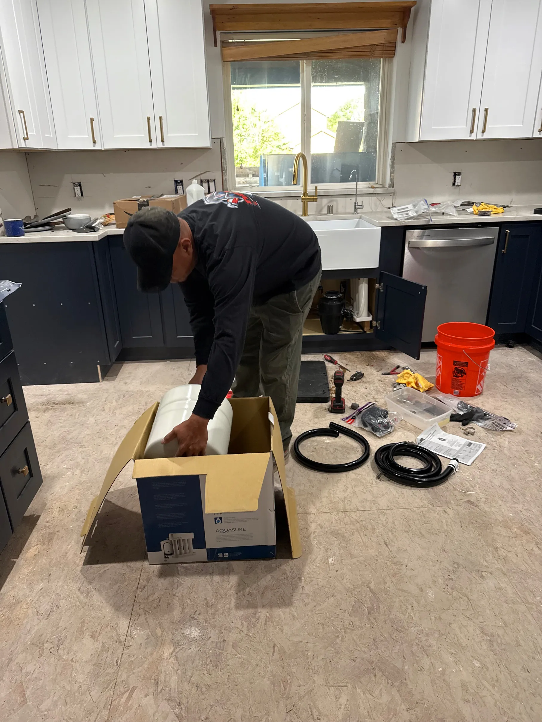 Plumber installing under-sink water filtration system in a kitchen with navy and white cabinets.