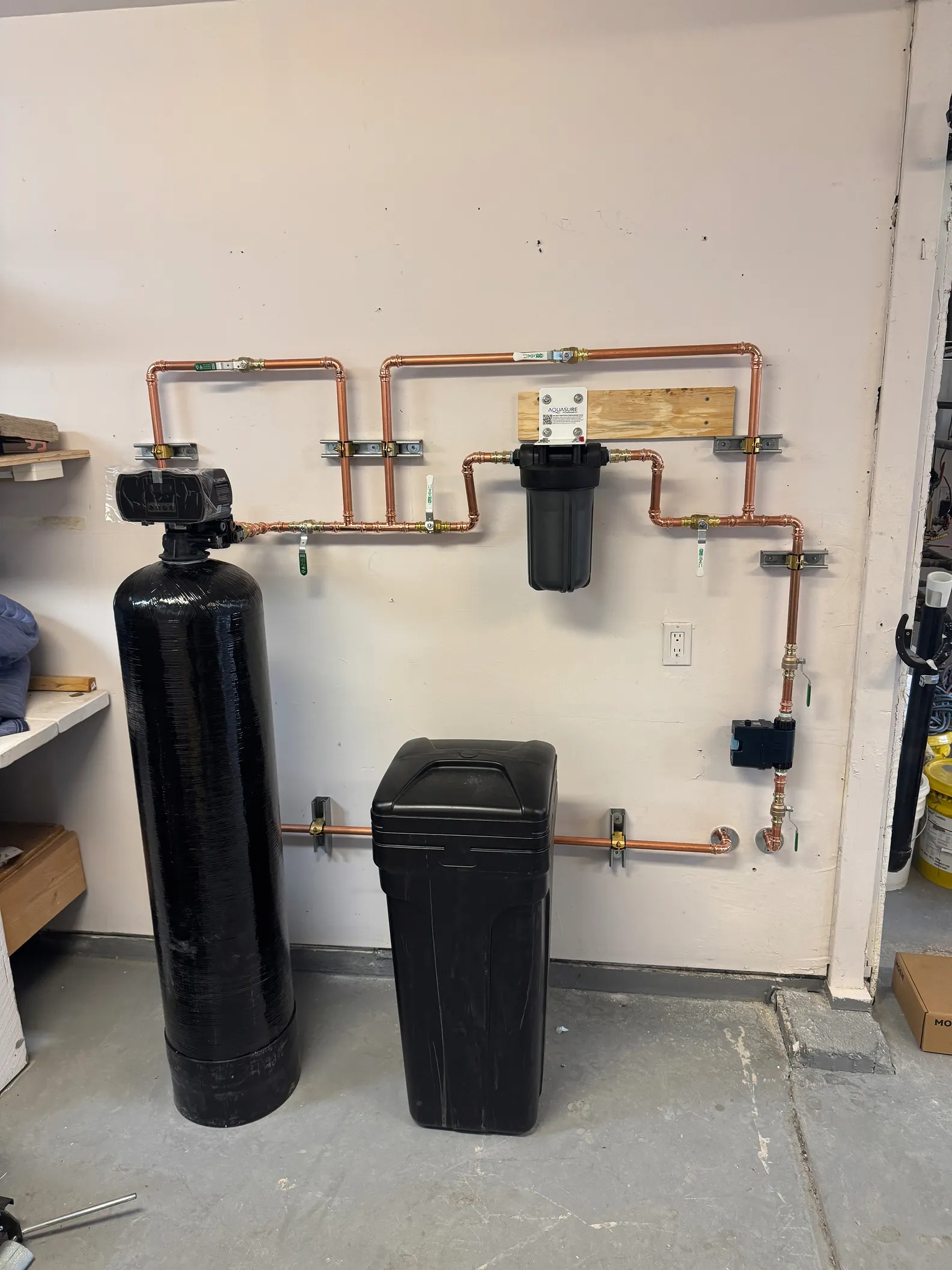 A residential water filtration system with black tanks and copper piping mounted on a beige wall in a utility room.