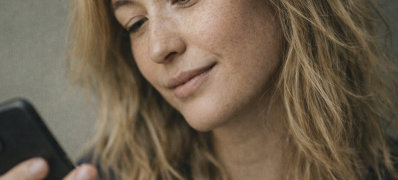 Närbild på en leende kvinna med långt, vågigt blont hår som tittar på en mobiltelefon.