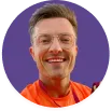 Smiling man with short hair and glasses wearing an orange shirt against a purple background.