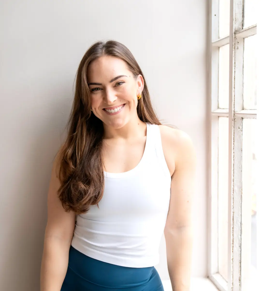 Smiling woman with long brown hair wearing a white tank top and blue leggings standing by a window.