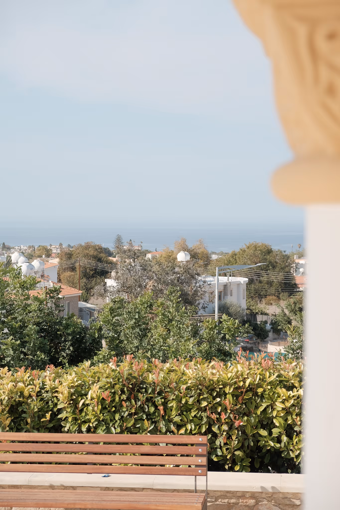 Wooden bench in front of dense green shrubbery with houses, trees, and a distant sea under a clear sky.