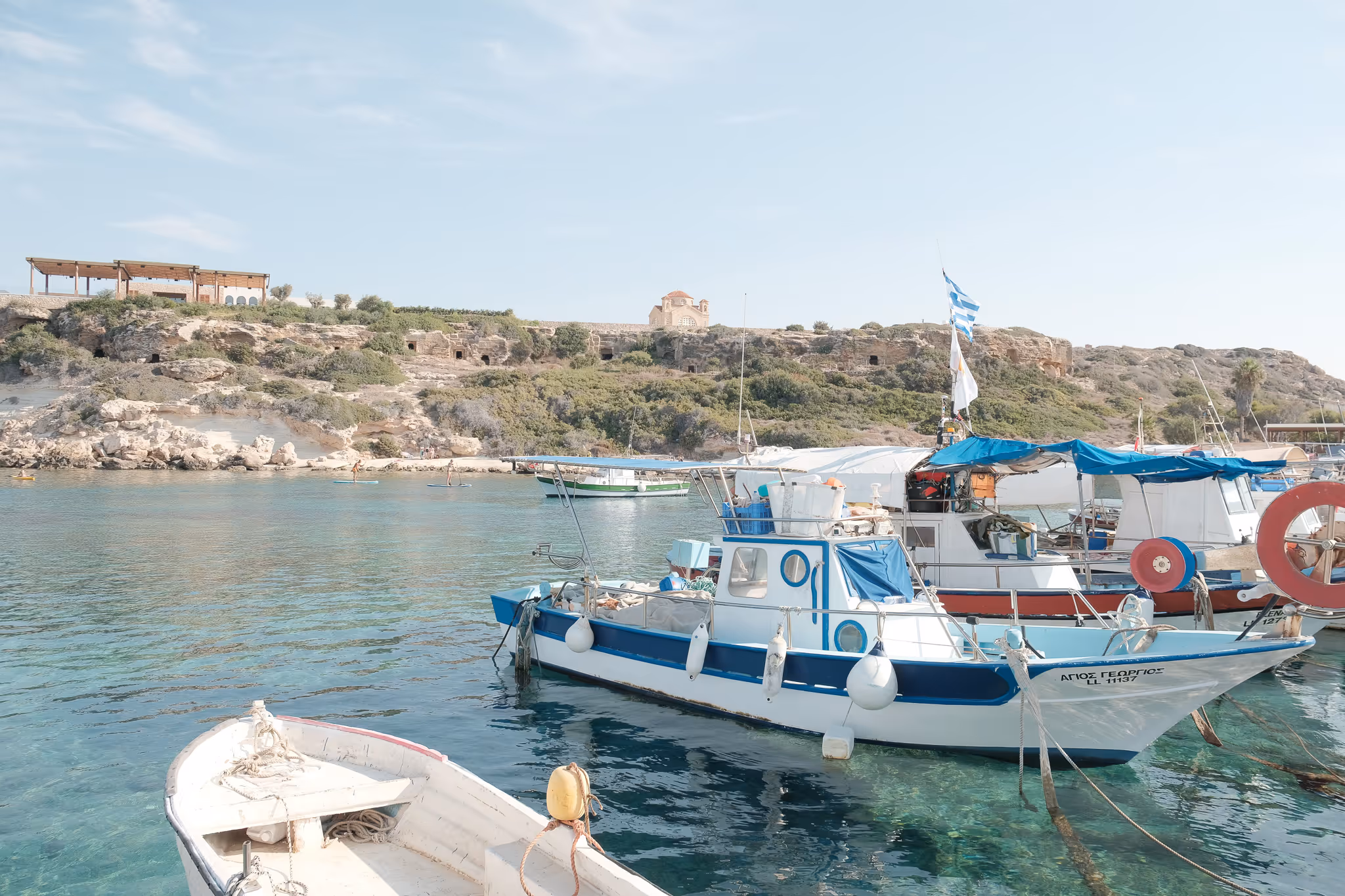 Small fishing boats with blue and white colors floating on clear turquoise water in a harbor with rocky cliffs and a small church in the background.
