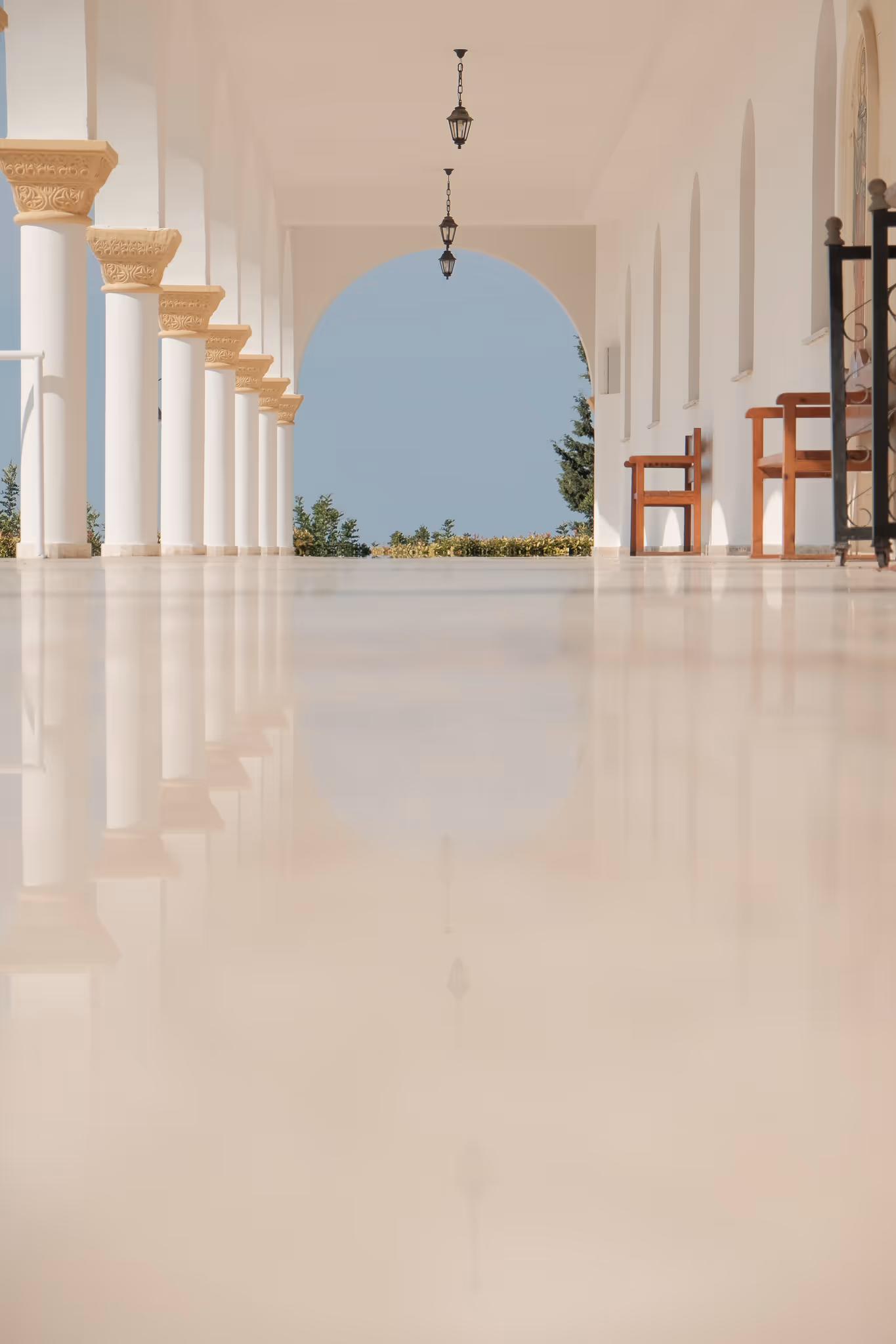 Bright corridor with white columns, hanging lanterns, wooden chairs, and a large archway opening to greenery outside.