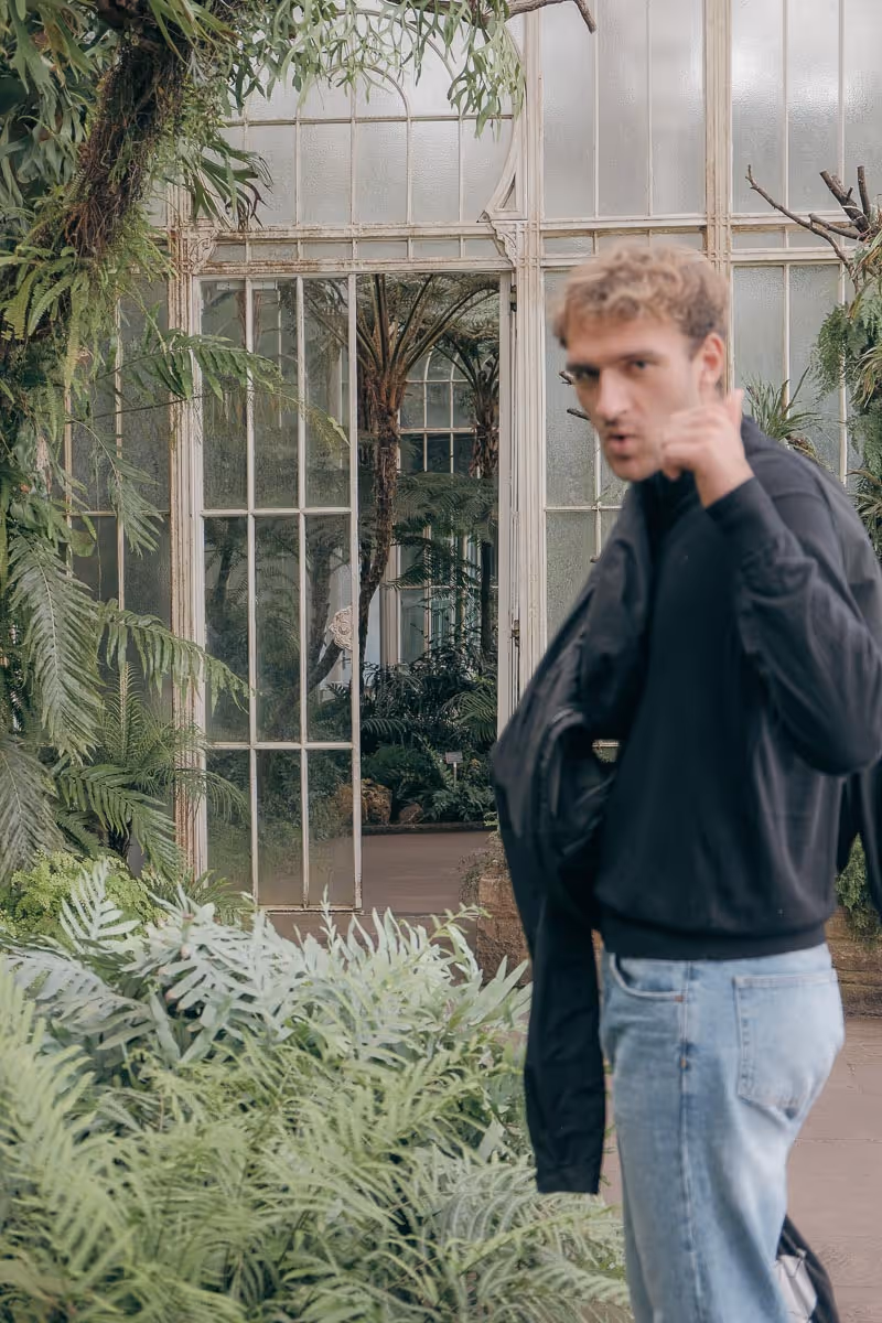 Blurred young man in black jacket and jeans pointing towards the camera in a greenhouse filled with lush green plants.