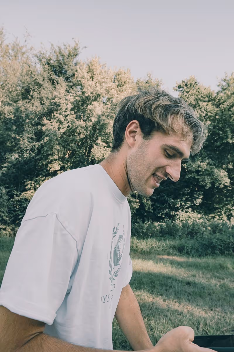 Young man in a white t-shirt smiling and looking down while standing outdoors with green trees in the background.