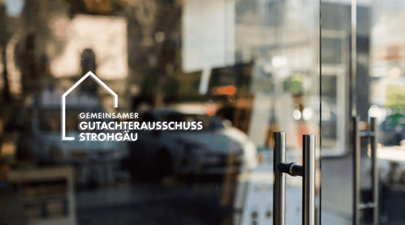 Glass door with a white outline of a house and text 'GEMEINSAMER GUTACHTERAUSSCHUSS STROHGÄU' reflected in the glass.
