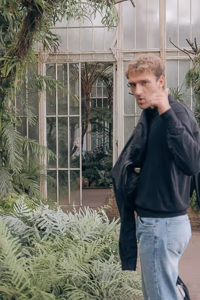 Blurred young man in black jacket and jeans pointing towards the camera in a greenhouse filled with lush green plants.