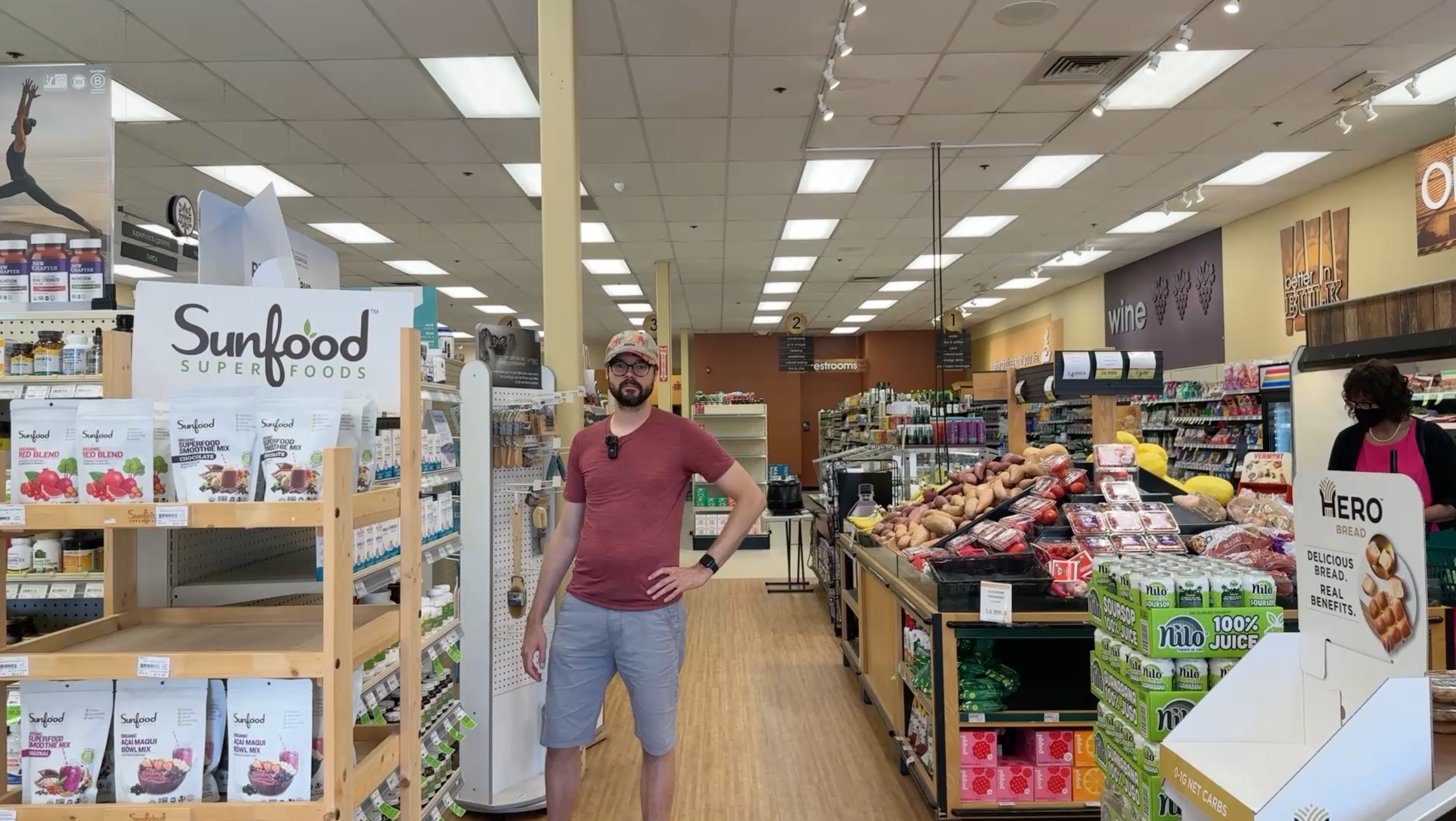 A man standing in a super market at better health store with fresh fruits and vegetable on the right side and other food items on the left