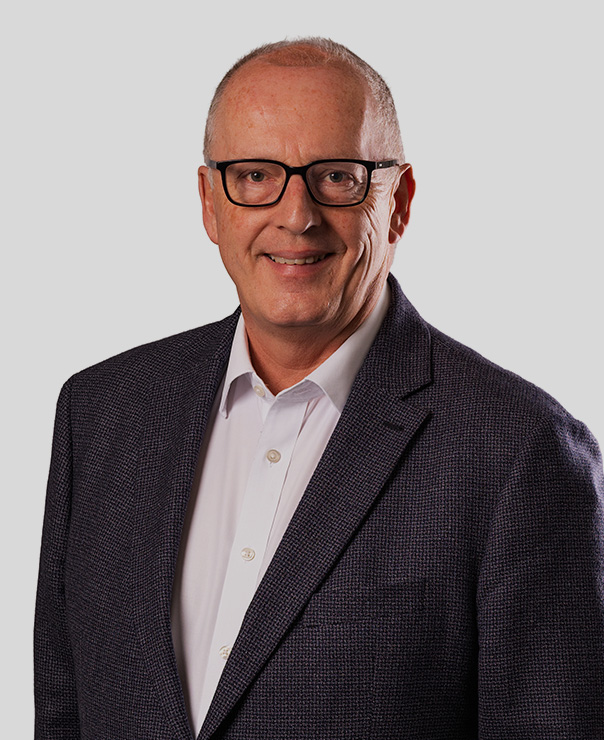 Smiling older man with glasses wearing a dark blazer and white shirt against a plain gray background.