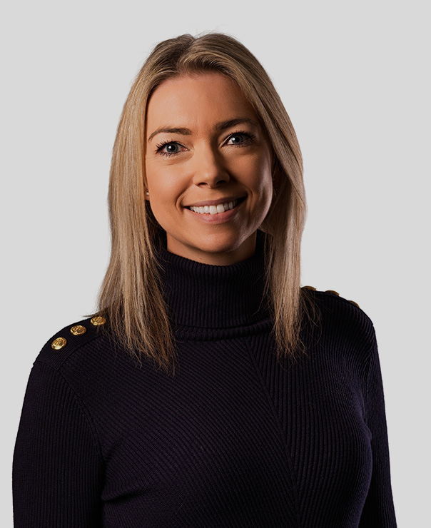 Smiling woman with straight blonde hair wearing a black turtleneck sweater with gold buttons on the shoulders.