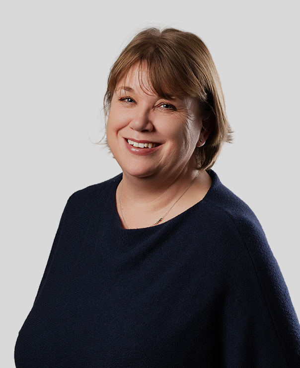 Smiling middle-aged woman with short brown hair wearing a dark navy sweater and necklace on light gray background.