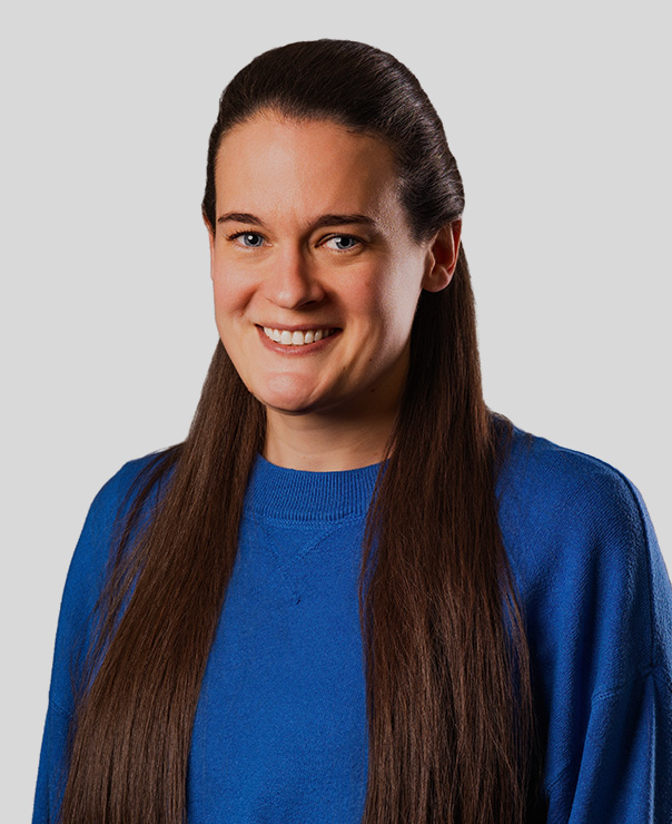 Smiling woman with long brown hair wearing a blue sweater against a gray background.