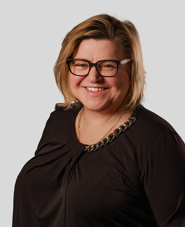 Smiling woman with short blond hair, wearing black-framed glasses and black top with chain detail.