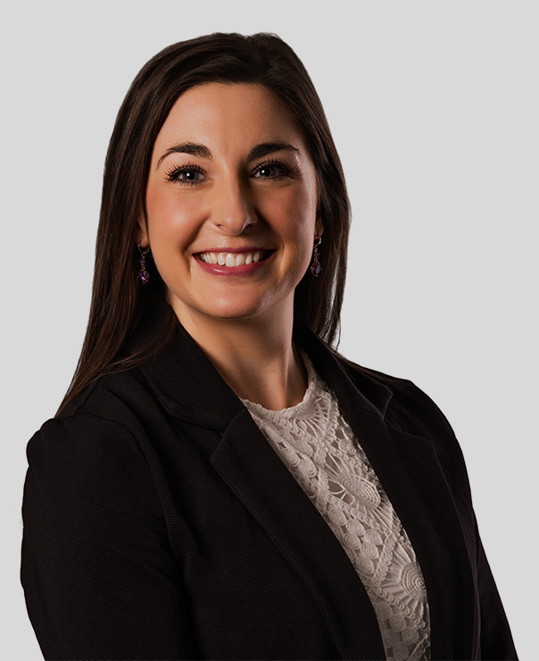 Smiling woman with long dark hair in a black blazer and white lace top against a gray background.