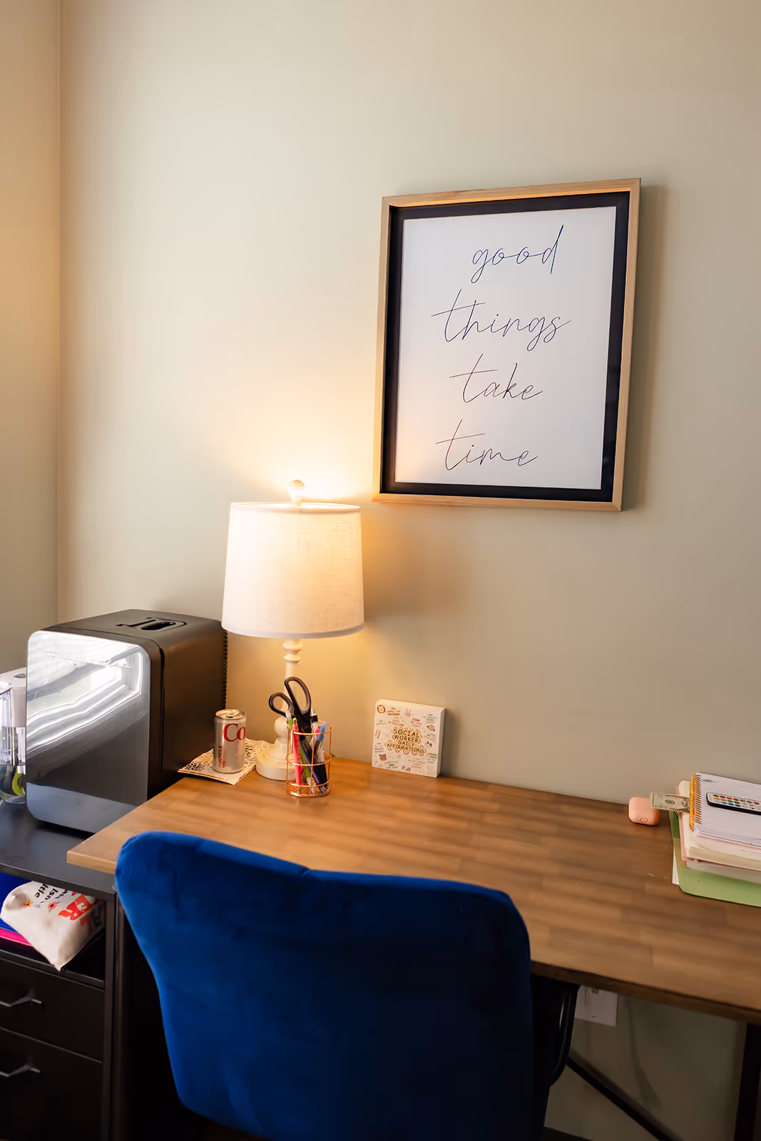 A wooden desk with a table lamp, a can of Diet Coke, stationery holder, and stacks of notebooks, with a blue chair in front and a framed sign on the wall saying 'good things take time'.