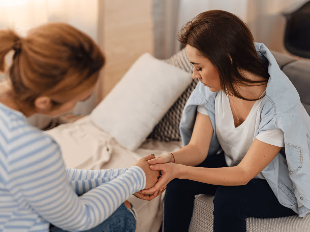 Two women sitting on a couch holding hands, one comforting the other who looks upset.