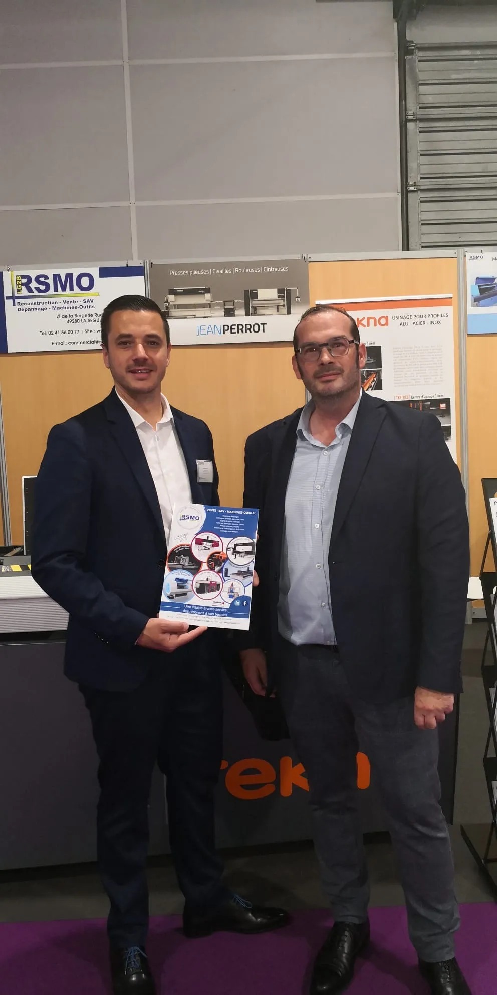 Deux hommes en costume souriants posant debout lors d'un salon professionnel, l'un tient une brochure de l'entreprise RSMO.