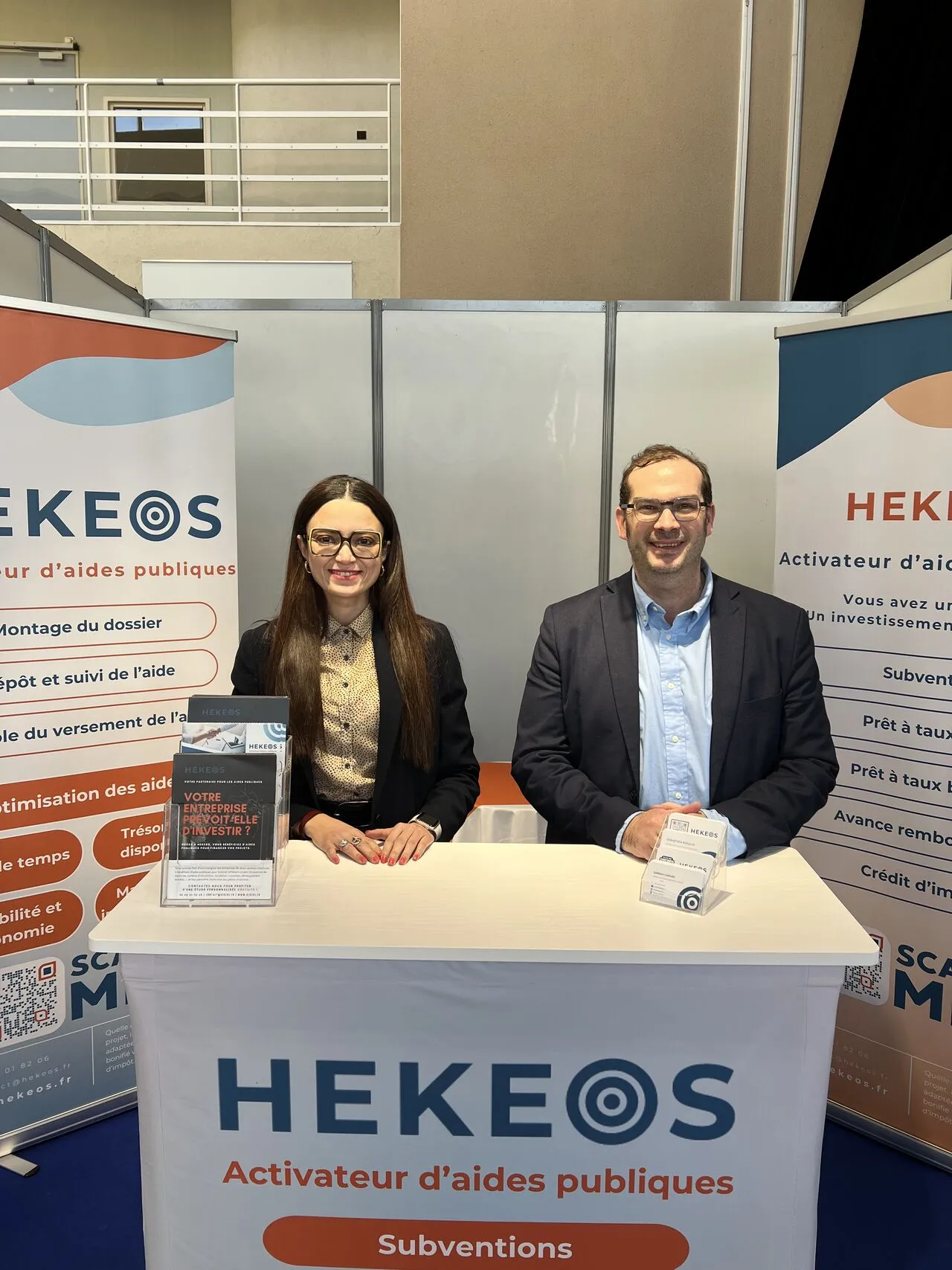 Deux personnes souriantes, un homme et une femme portant des lunettes, posent derrière un comptoir avec la bannière HEKEOS, activateur d’aides publiques.