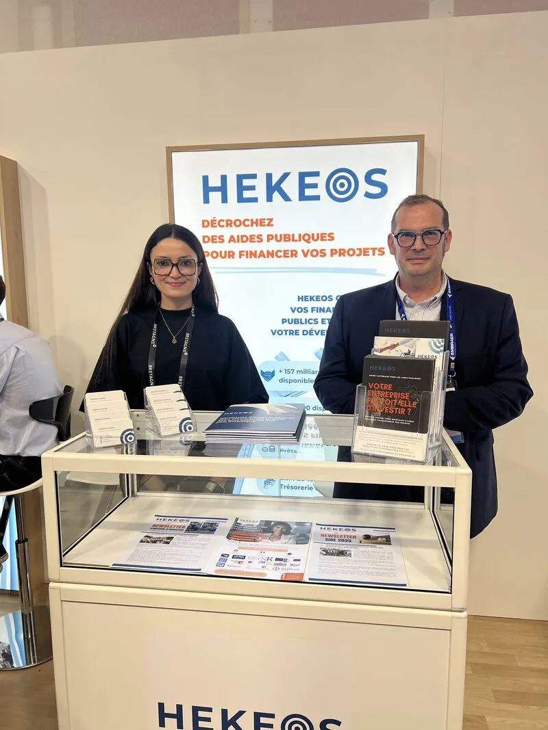 Deux personnes souriantes, homme et femme, debout derrière un comptoir d'exposition avec des brochures HEKEOS pour le financement public de projets.