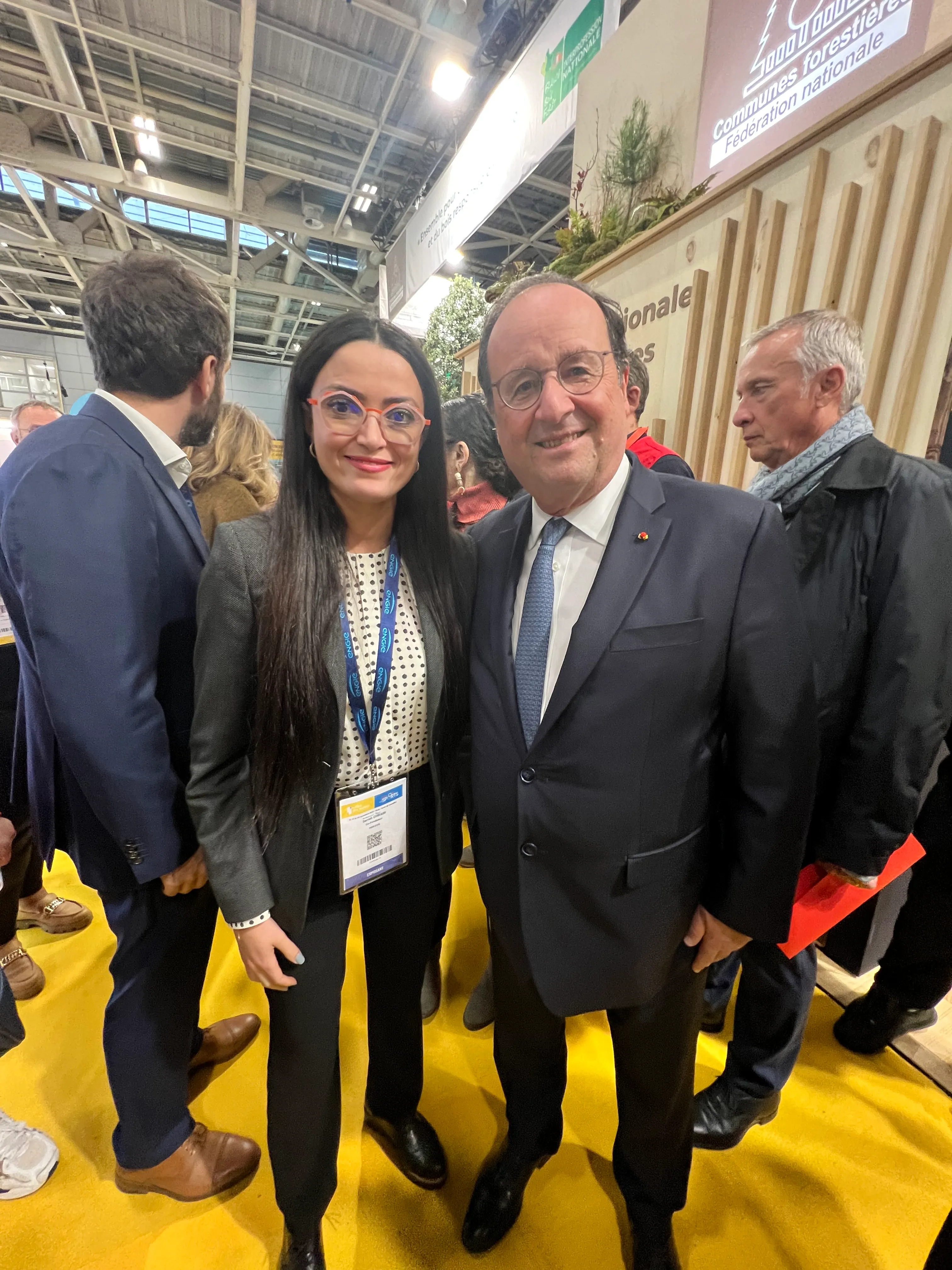 Une femme en tenue professionnelle posant à côté d'un homme en costume lors d'un événement intérieur avec des personnes en arrière-plan.