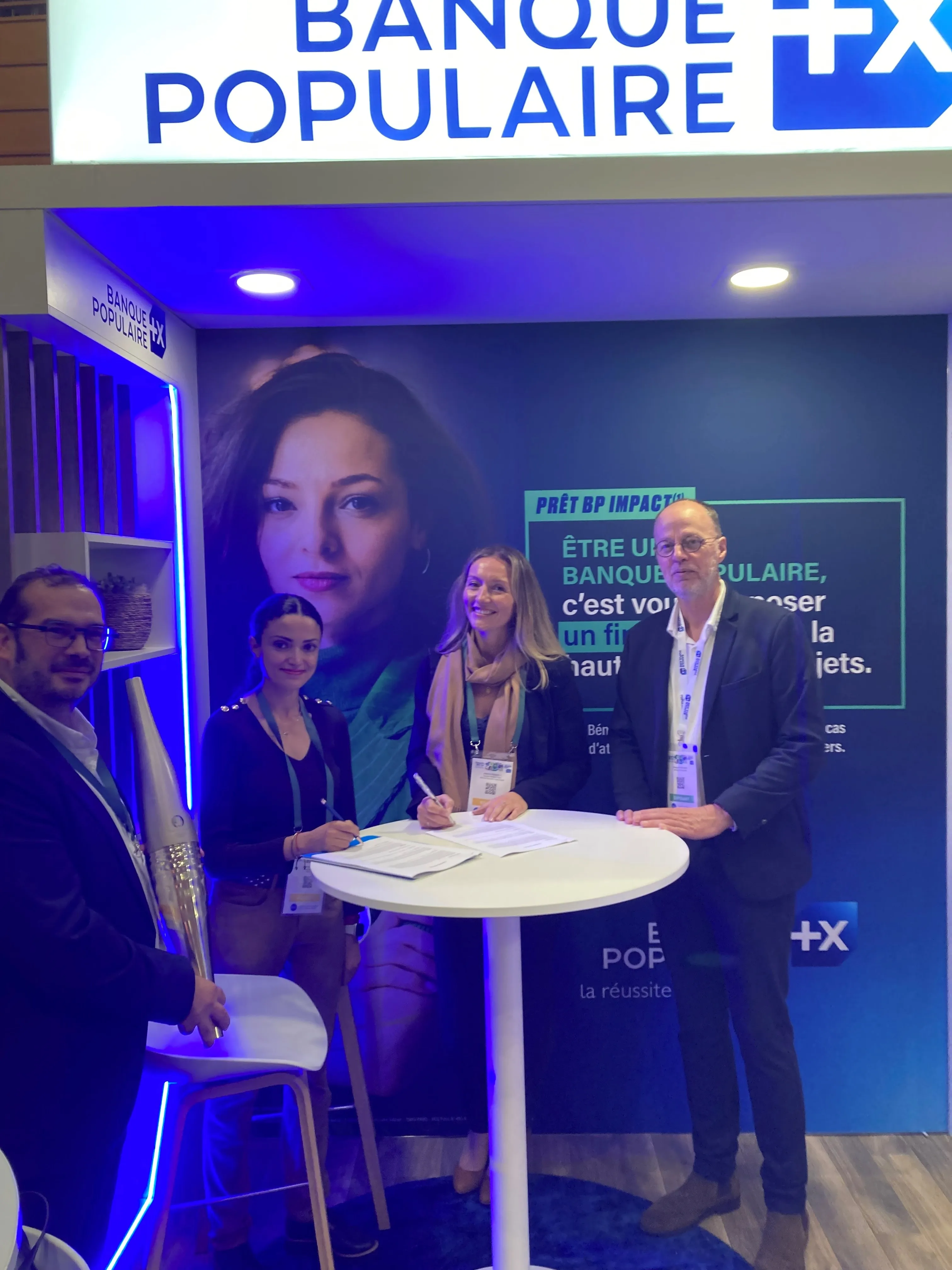 Quatre personnes souriantes debout autour d'une table haute lors d'un événement au stand de la Banque Populaire, avec un grand panneau bleu et blanc en arrière-plan.