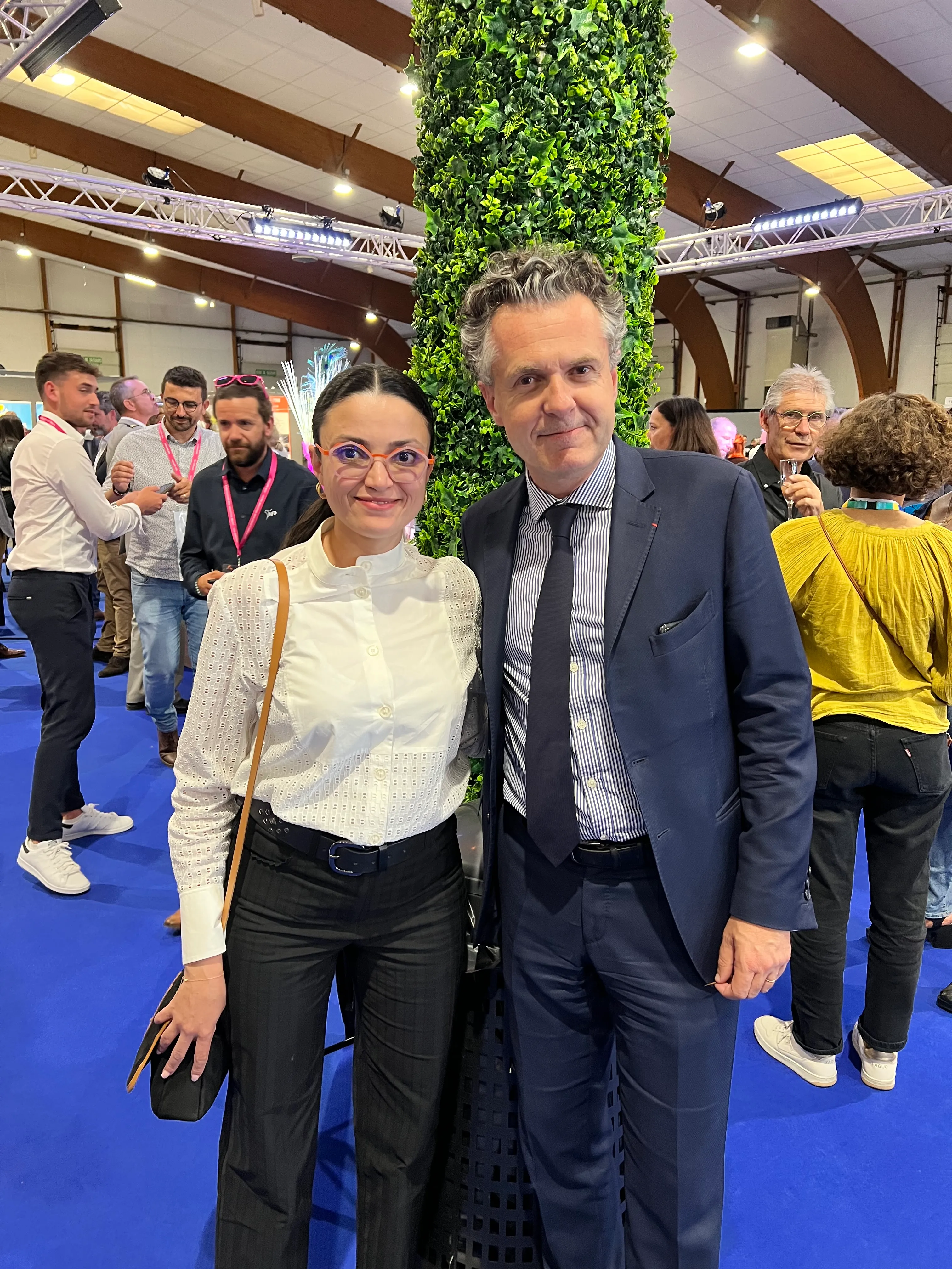 Un homme et une femme posent ensemble devant une colonne couverte de verdure lors d'un événement à l'intérieur avec plusieurs personnes en arrière-plan.
