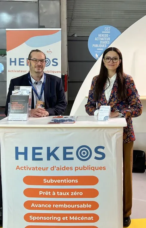 Deux personnes souriantes debout derrière un comptoir avec une bannière HEKEOS, activateur d'aides publiques, affichant des services comme subventions, prêt à taux zéro, avance remboursable, et sponsoring et mécénat.