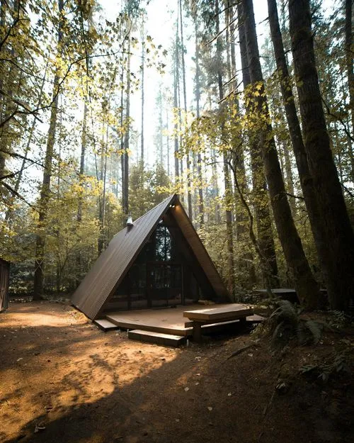 A modern A-frame cabin nestled in a sun-dappled forest, surrounded by tall trees and autumn foliage, with a wooden deck in front.