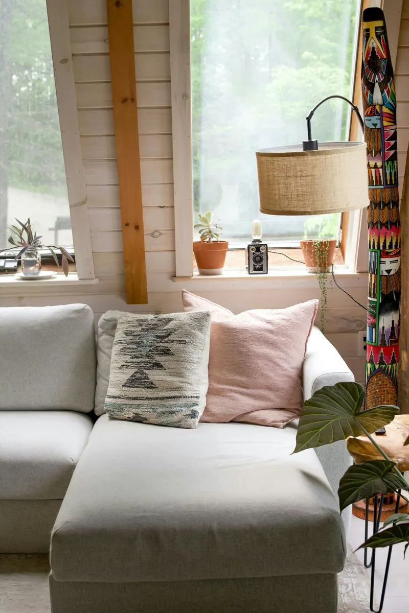 Cozy living room corner with a light gray couch adorned with decorative pillows, natural light filtering through windows, and potted plants.