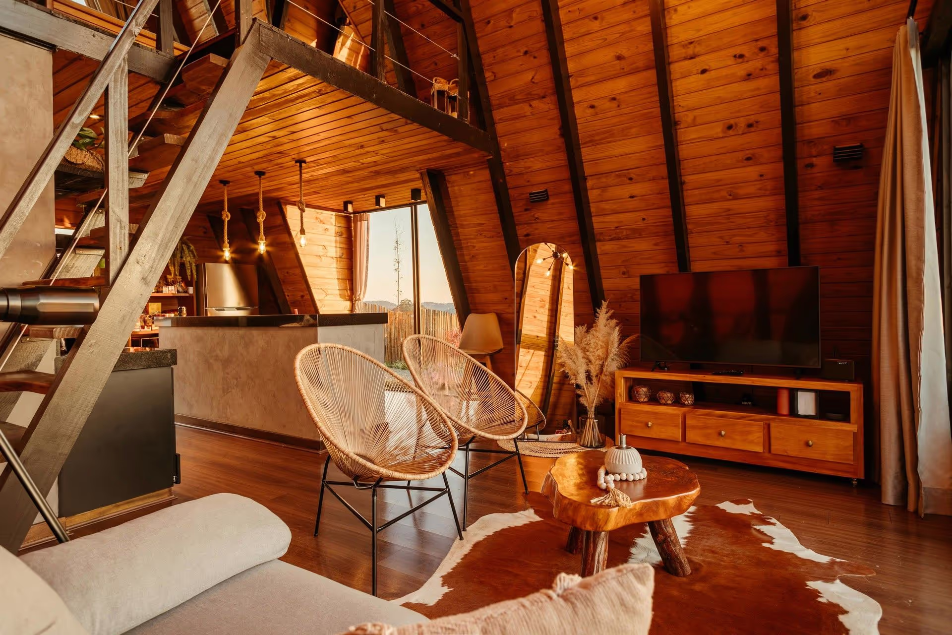 Cozy, wooden-finished interior of an A-frame cabin, featuring a spiral staircase, comfortable seating, and warm lighting.