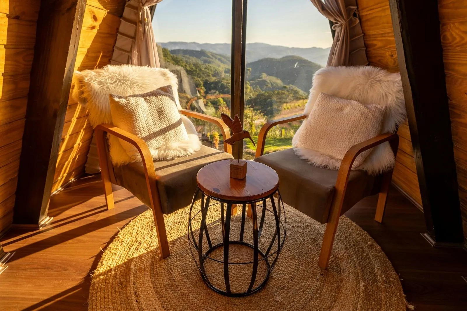 Cozy seating area with two plush chairs and a round table, overlooking scenic mountains through a large window. Natural light enhances warmth.