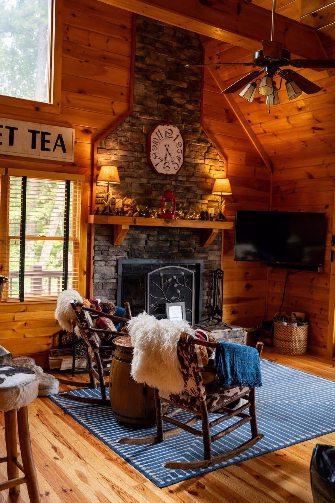 Cozy wooden cabin interior featuring a stone fireplace, rustic armchairs, a blue rug, and a wall clock, creating a warm atmosphere.