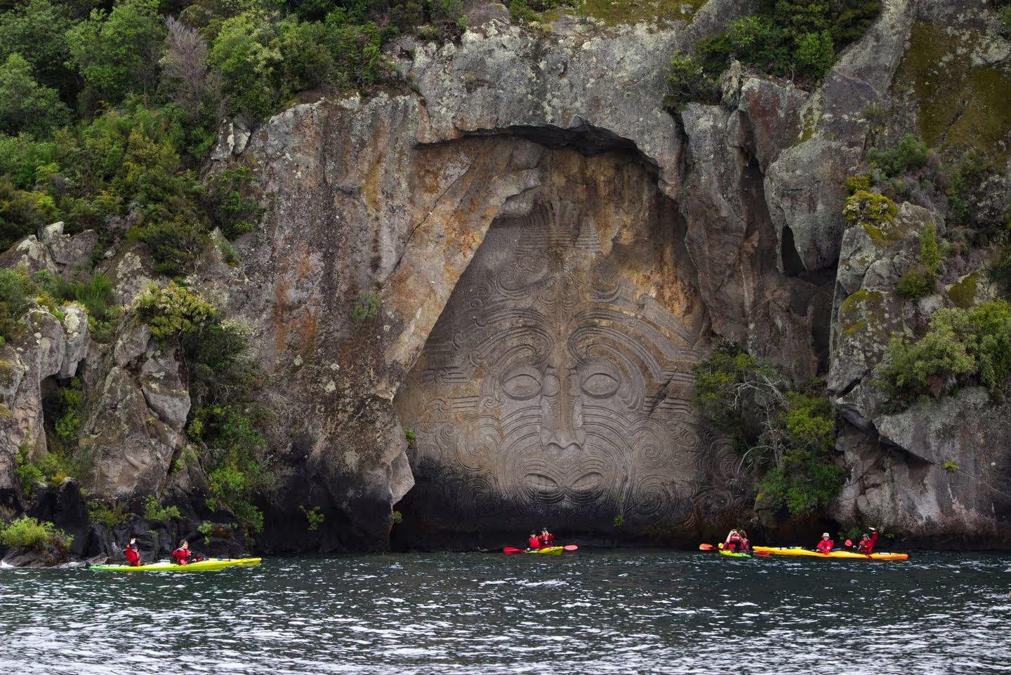 Kayakers paddle in the tranquil waters near a large, eroded rock formation, surrounded by lush greenery and cliffs.