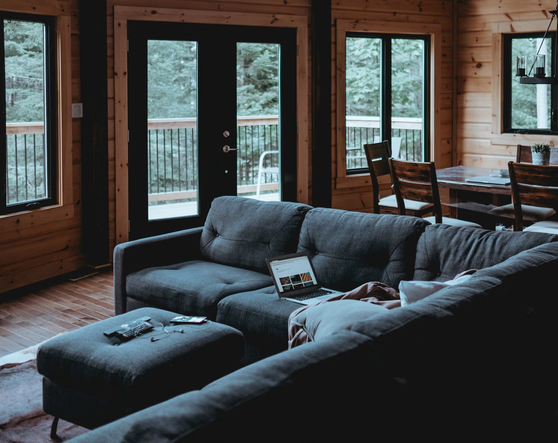 Cozy living room with a gray sectional sofa, a laptop open on the seat, and large windows overlooking a green forest.